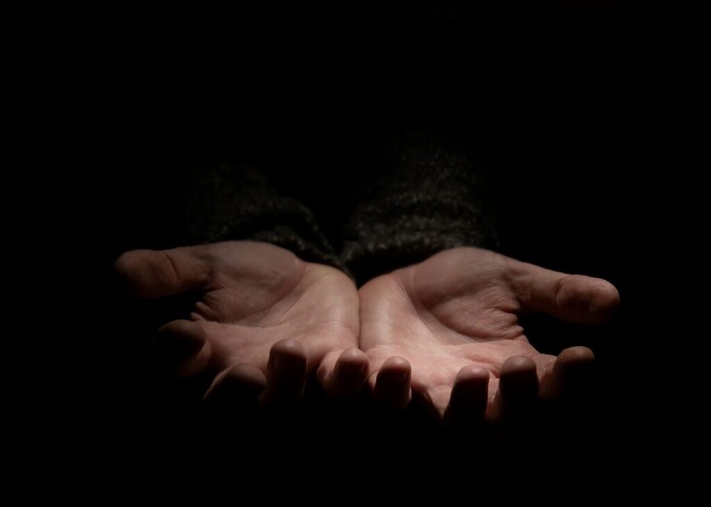 Two open hands with palms facing up are lit by a dim light, emerging from a dark background.