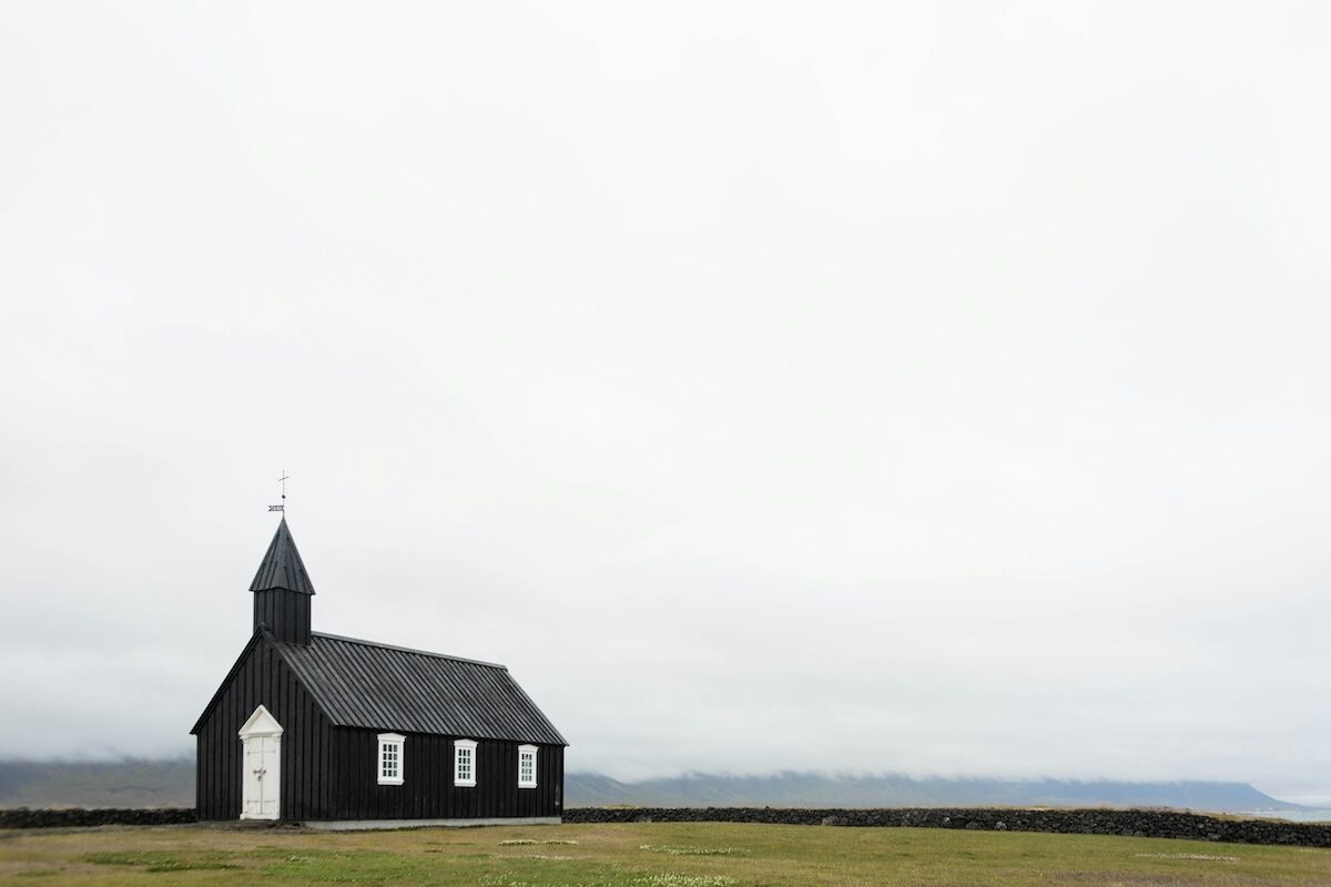 A small black church with a white door stands alone on a grassy field under an overcast sky.