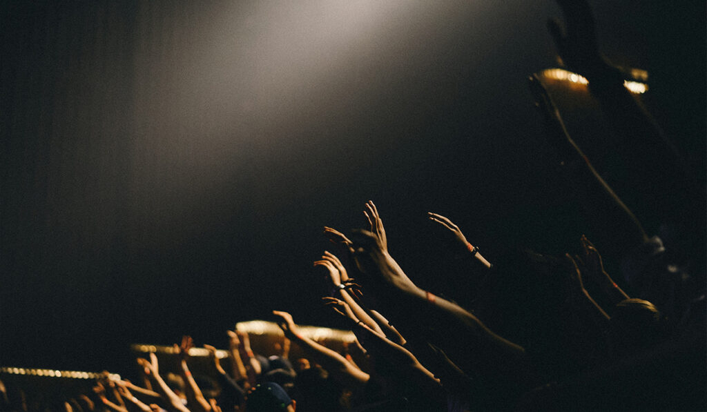 Hands are held up high in worship in a dark auditorium.