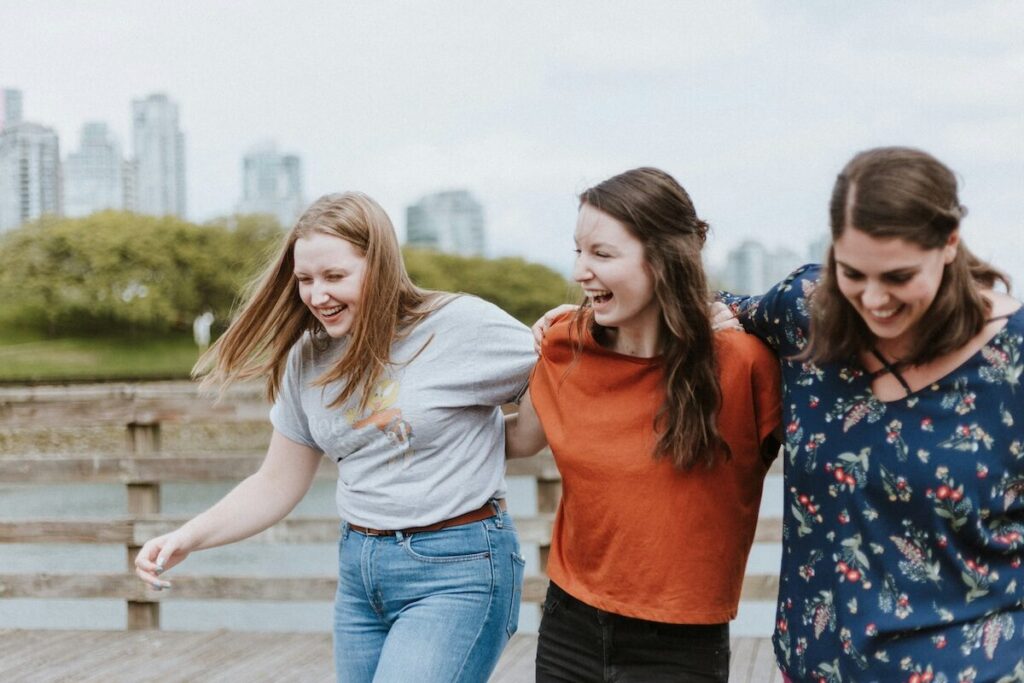 Three young women walk side by side on a wooden boardwalk outdoors, smiling and laughing, with a cityscape and greenery in the background.