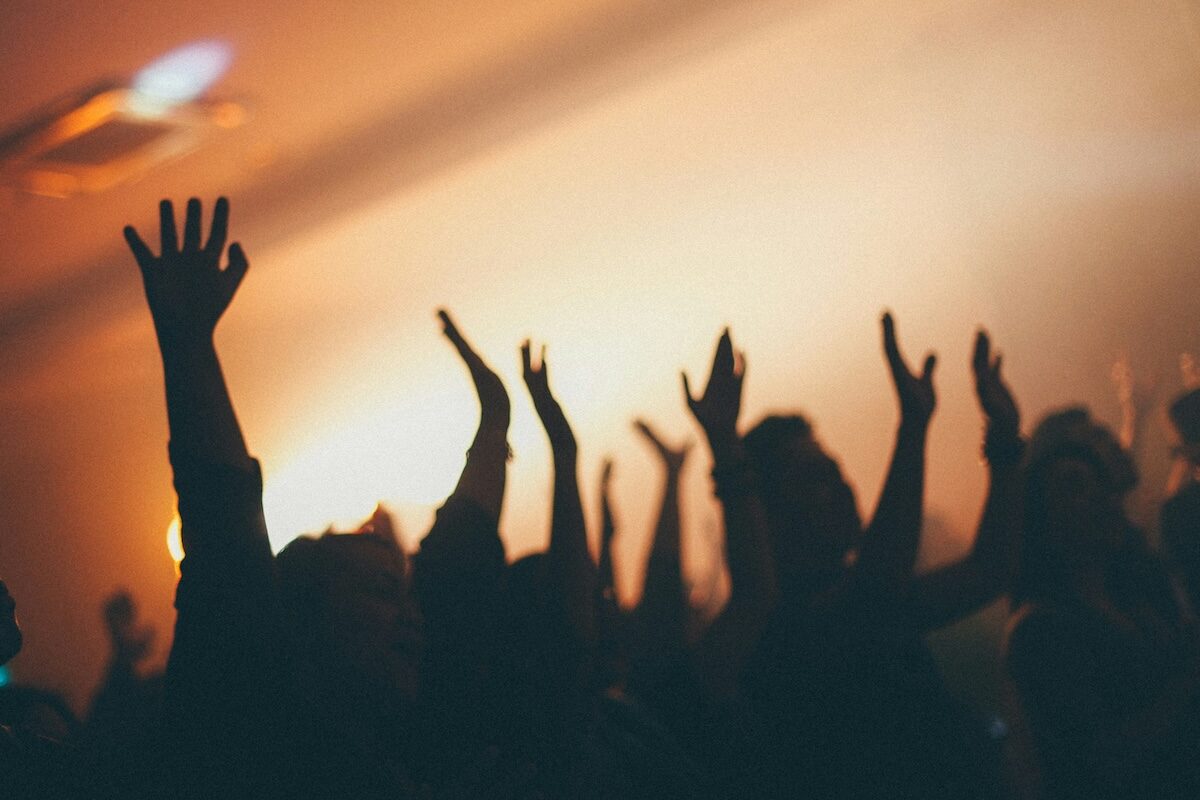 Silhouetted people raise their hands in the air at a crowded indoor event with warm lighting in the background.