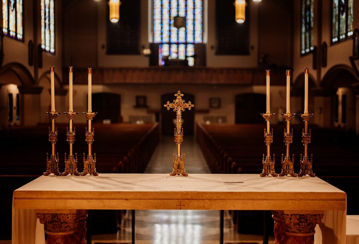 An empty church interior with a marble altar in the foreground, featuring a cross and two sets of three unlit candles on ornate holders.