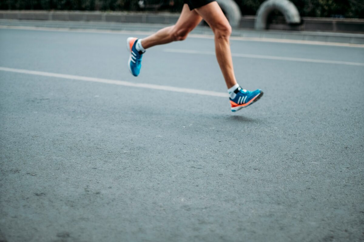 A person running on a paved road, wearing blue athletic shoes, black shorts, and a dark top. Only the legs are visible in the image.