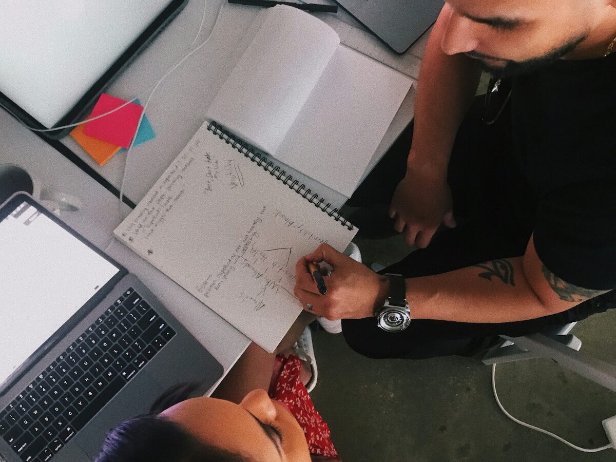 Two people sit at a desk with a laptop, notebooks, and sticky notes, collaborating and taking notes on a spiral notebook.