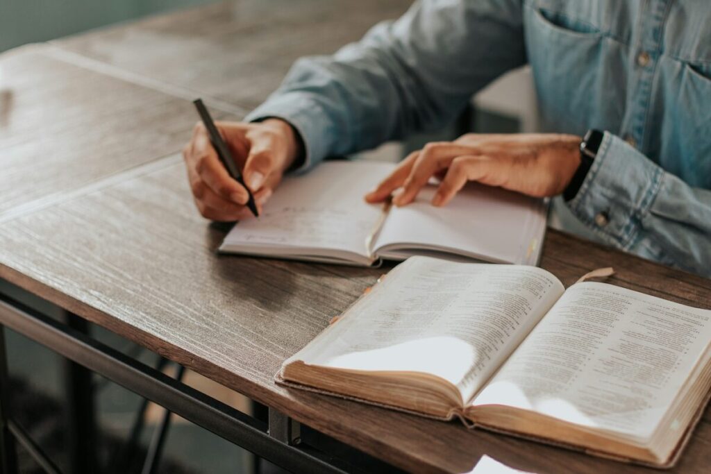 A person wearing a denim shirt writes in a notebook at a desk, with an open book placed nearby.