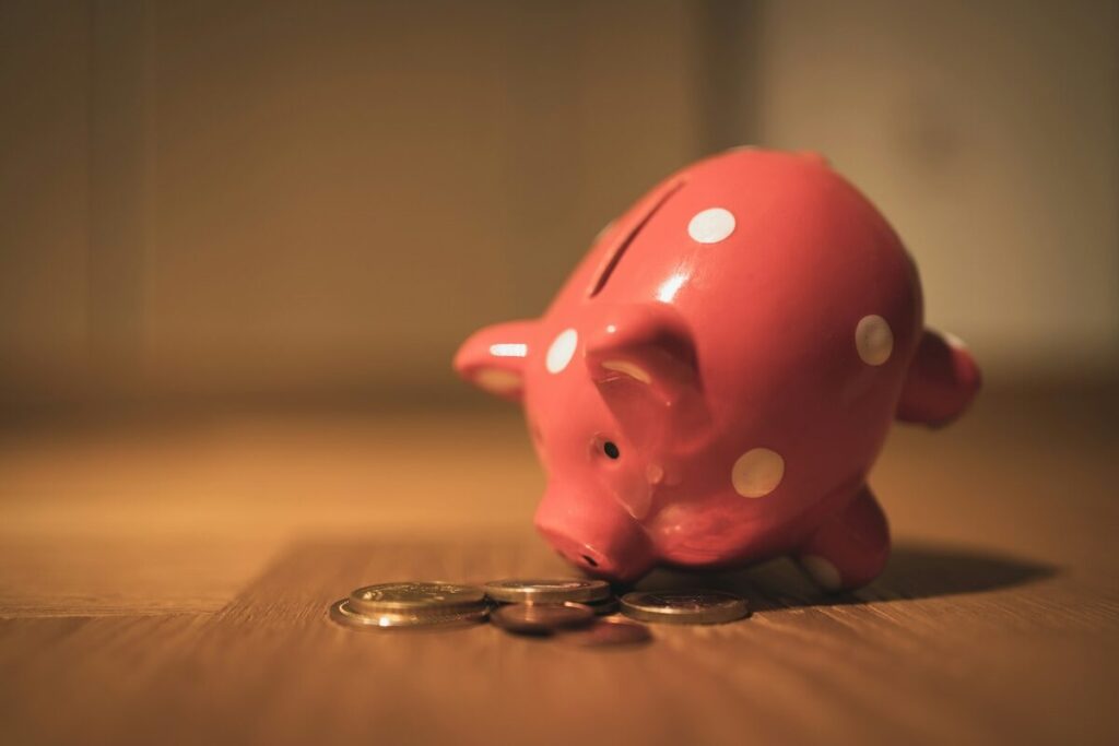 A pink piggy bank with white spots is tipped over on a wooden surface, with coins scattered in front of it.