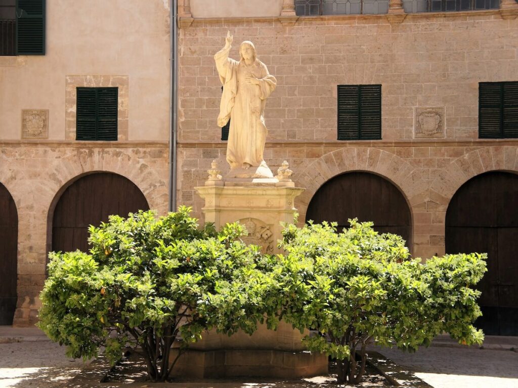 A stone statue of a robed figure stands on a pedestal behind green shrubs, set against a historic building with arched doorways and shuttered windows.