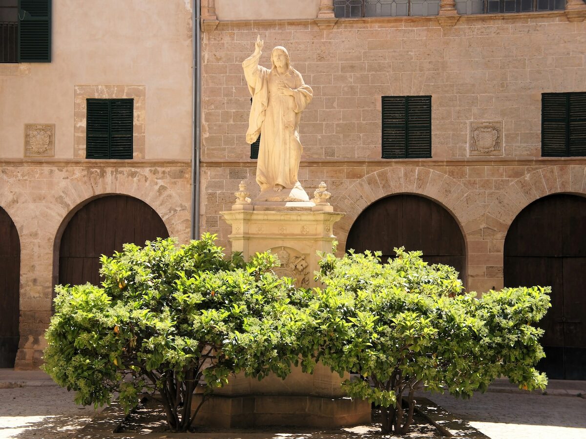 A stone statue of a robed figure stands on a pedestal behind green shrubs, set against a historic building with arched doorways and shuttered windows.