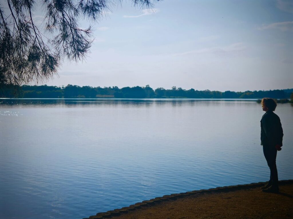 A person stands on the edge of a calm lake, looking out over the water with trees and a cloudy sky in the background.
