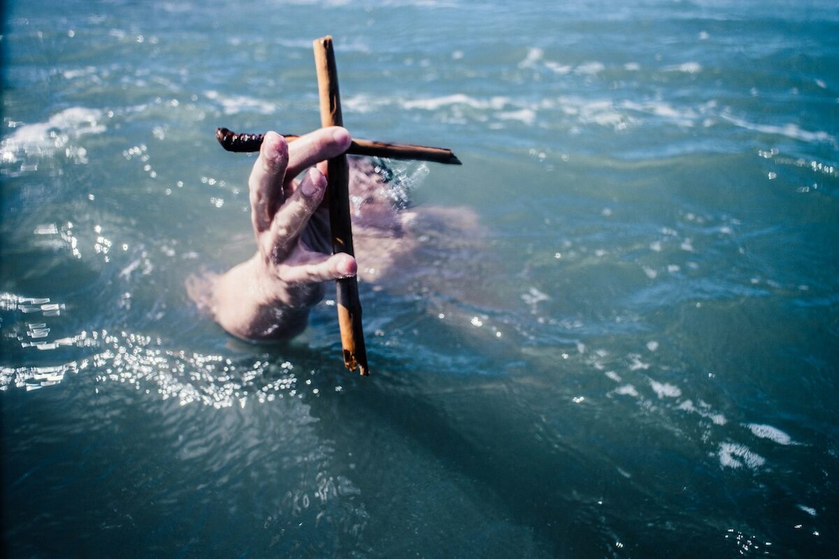A hand holding a wooden cross emerges from the surface of a body of water, with waves surrounding the hand.