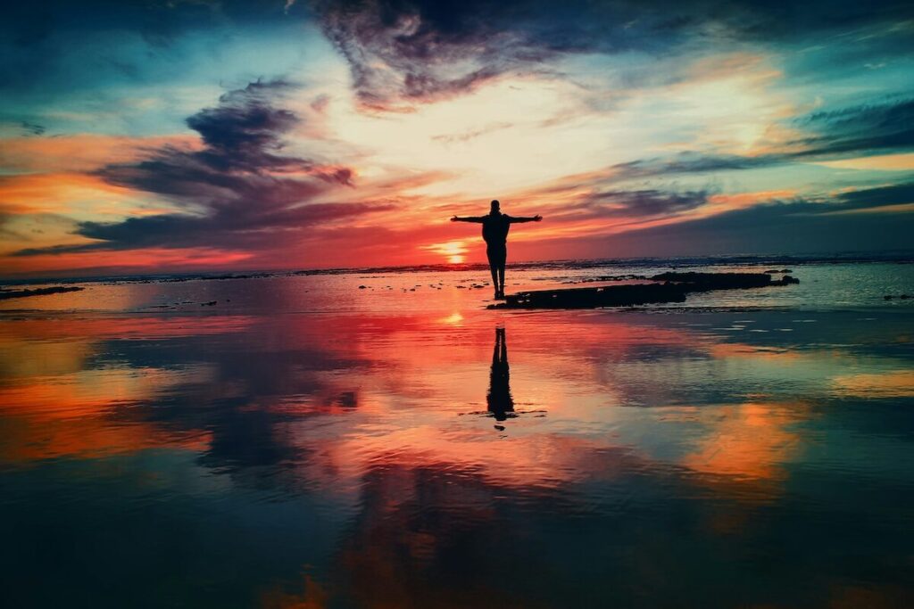 A person stands with arms outstretched on a shoreline at sunset, reflected in calm water with vibrant clouds and colors in the sky.