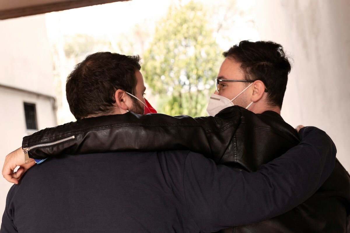 Two people wearing face masks and jackets stand close together with their arms around each other, facing away from the camera.