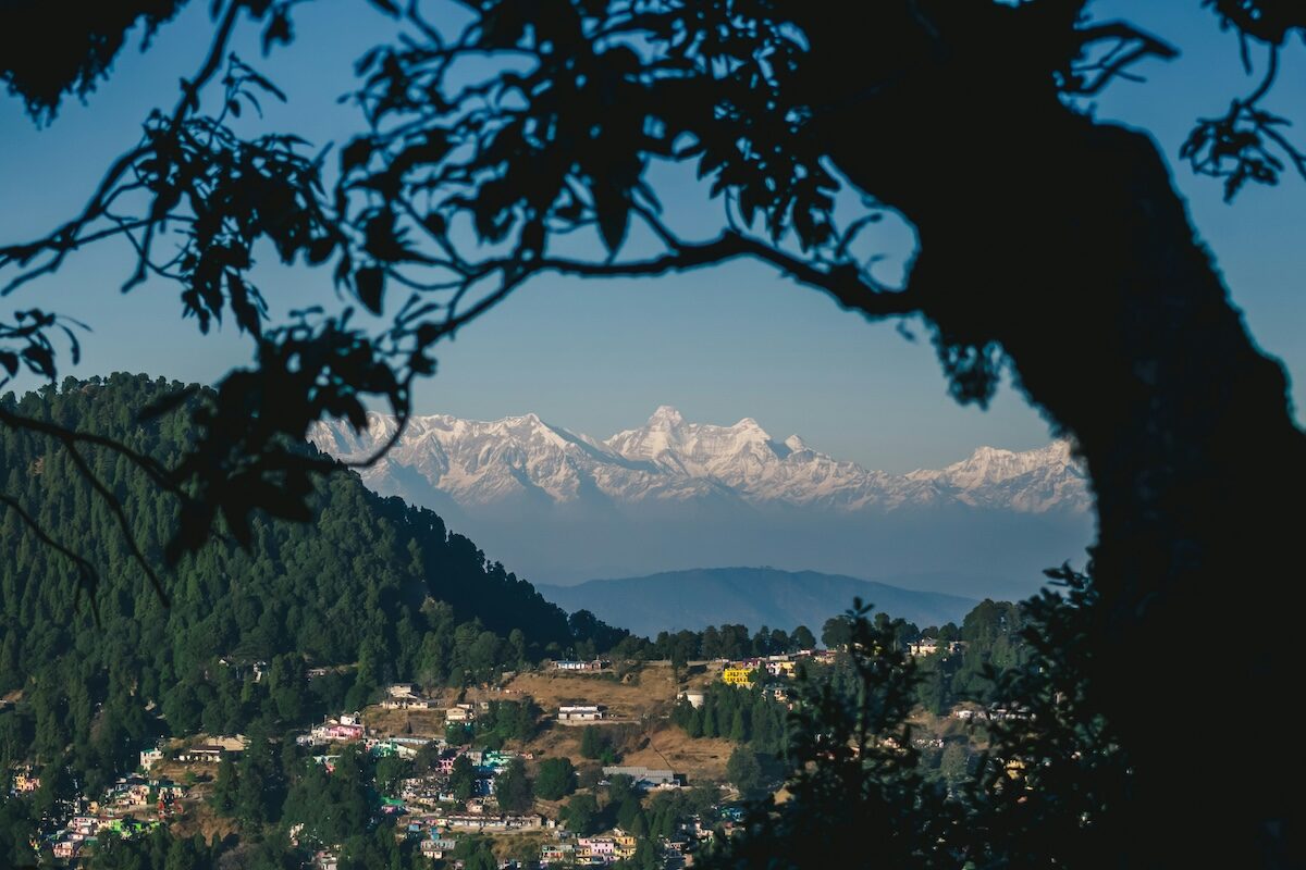 Mountain village with scattered houses in the foreground, framed by tree branches, with snow-capped peaks visible in the distant background under a clear blue sky.
