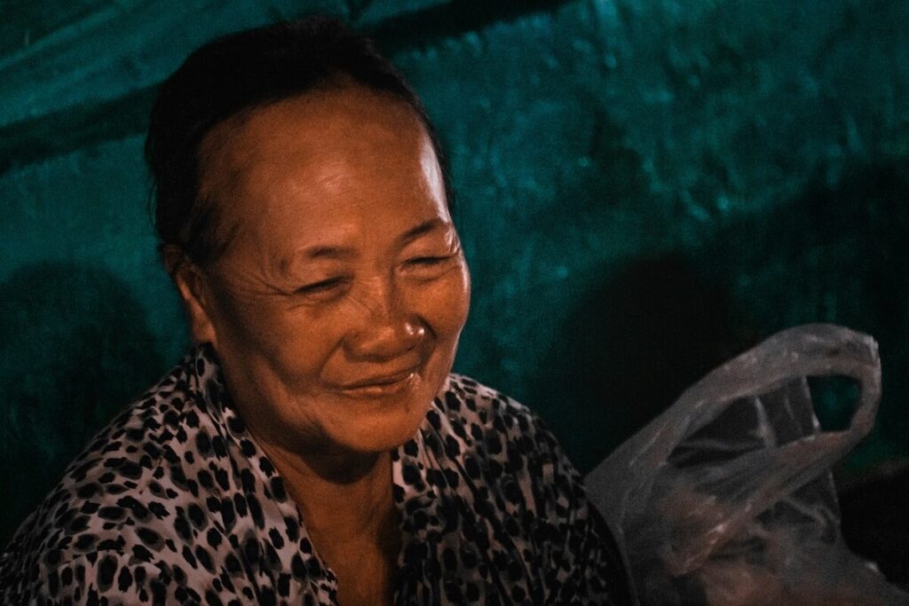 An older woman with short hair smiles softly, wearing a patterned shirt, with a plastic bag and a green background behind her.