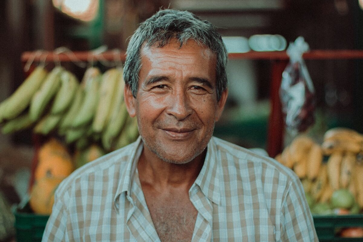 Older man with gray hair and plaid shirt stands in front of a market stall with bananas and produce in the background.