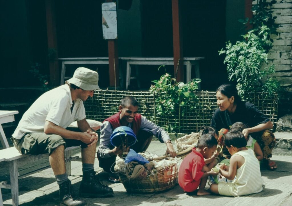 A group of adults and children sit together outdoors on a sunny day, some sitting on the ground and one adult sitting on a bench, talking and interacting near baskets.
