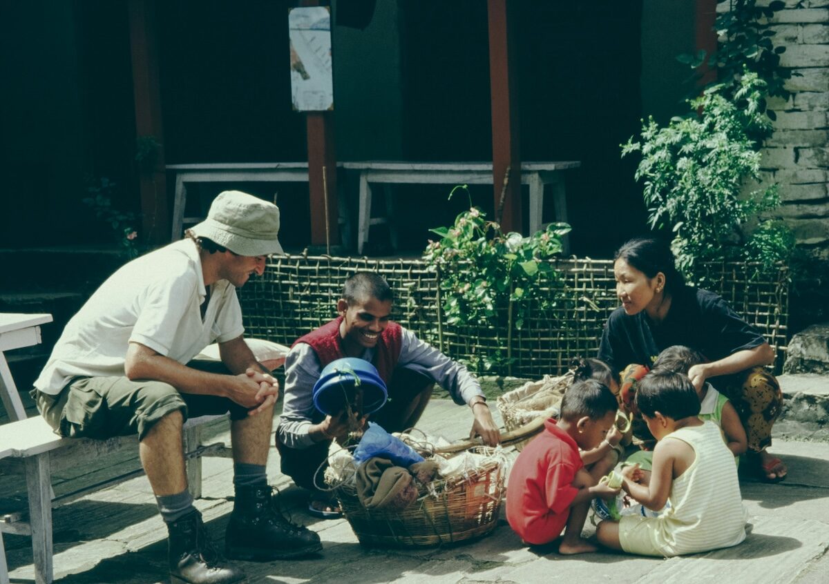 A group of adults and children sit together outdoors on a sunny day, some sitting on the ground and one adult sitting on a bench, talking and interacting near baskets.