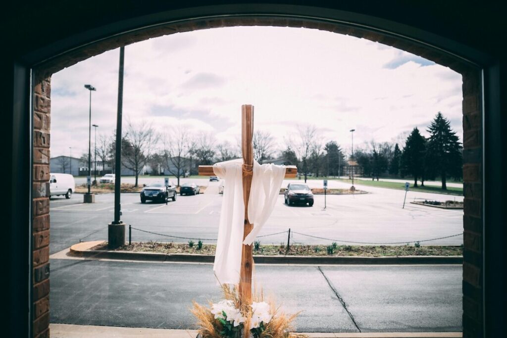 A wooden cross draped with a white cloth stands at the entrance of a building, overlooking a parking lot and grassy area on a cloudy day.