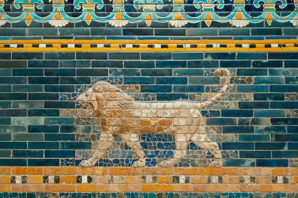 Ancient mosaic of a roaring lion in profile, created with blue, yellow, and white tiles, on a decorative brick wall.