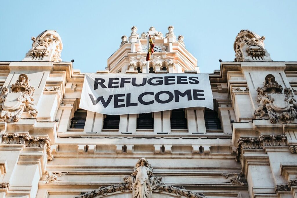 A large banner reading REFUGEES WELCOME hangs on the facade of an ornate historic building under a clear sky.