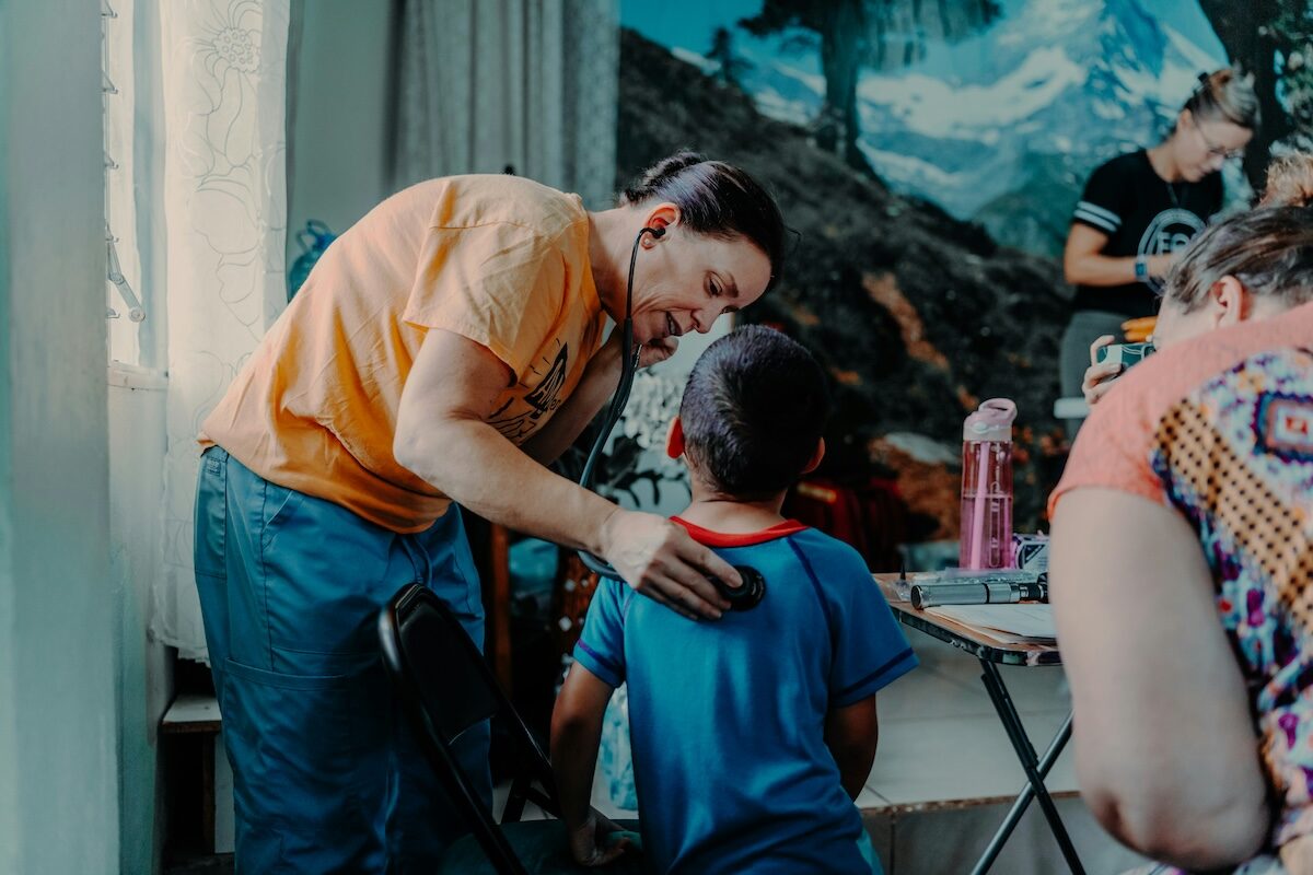 An adult gently places a hand on a childs back while talking to them indoors, with other people and a mountain mural visible in the background.