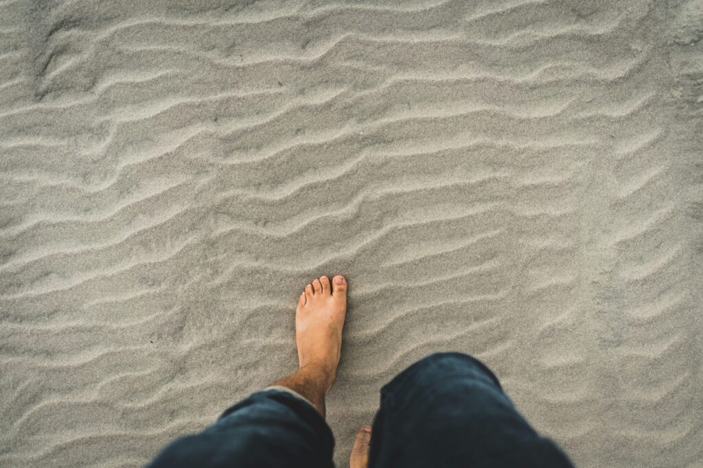 A person with bare feet and wearing shorts stands on rippled sand.