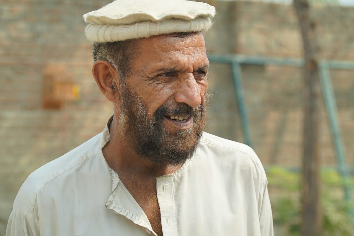 A middle-aged man with a beard wearing a traditional light-colored cap and shirt stands outdoors near a brick wall.