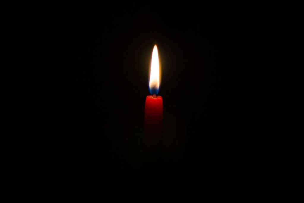 A single red candle with a bright flame burns in darkness, illuminating its immediate surroundings.