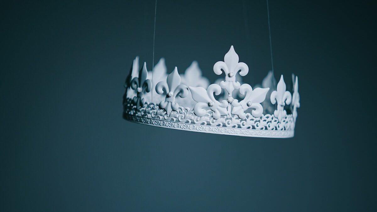 A white ornate crown with fleur-de-lis details hangs suspended by thin strings against a plain, dark background.
