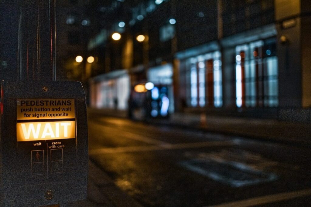 A pedestrian crossing button displays an illuminated WAIT sign at night on a city street with blurred buildings and lights in the background.