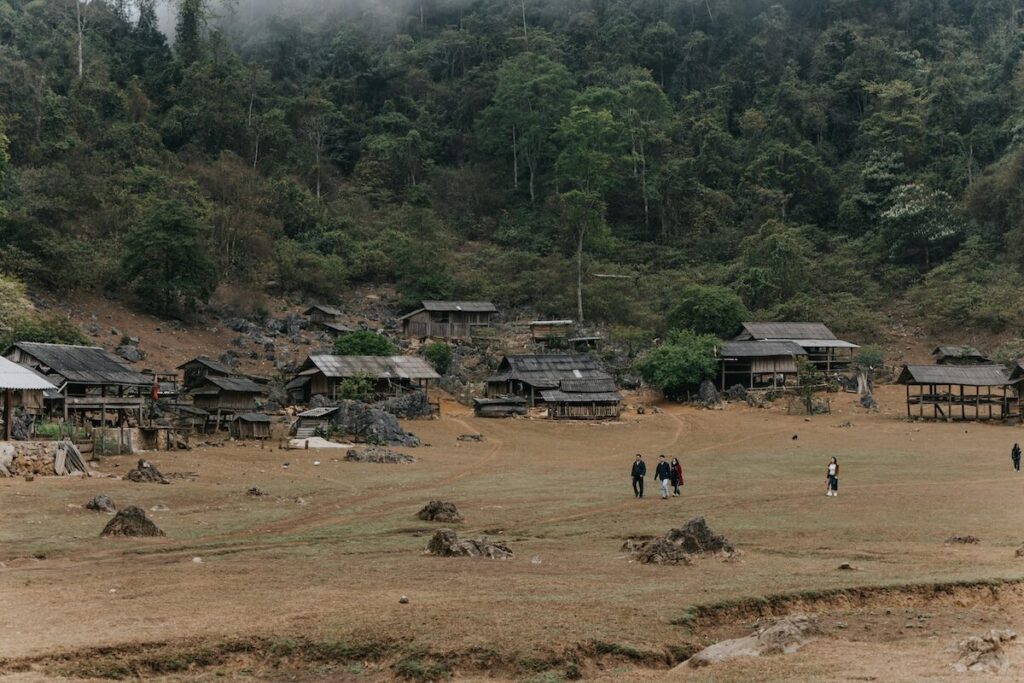 A rural village with wooden houses is surrounded by trees and hills, with a few people walking across an open, grassy area in the foreground.