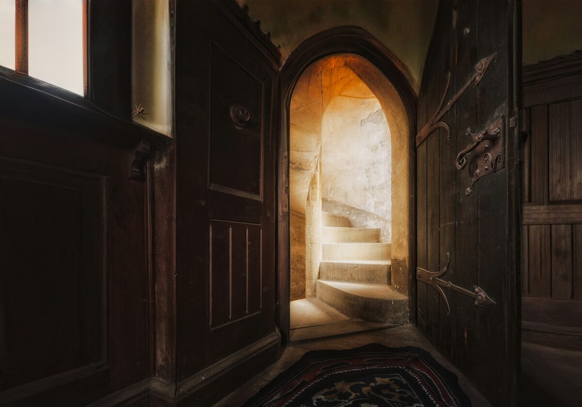 A dimly lit interior with a wooden door open to reveal a narrow stone spiral staircase illuminated by sunlight from above.