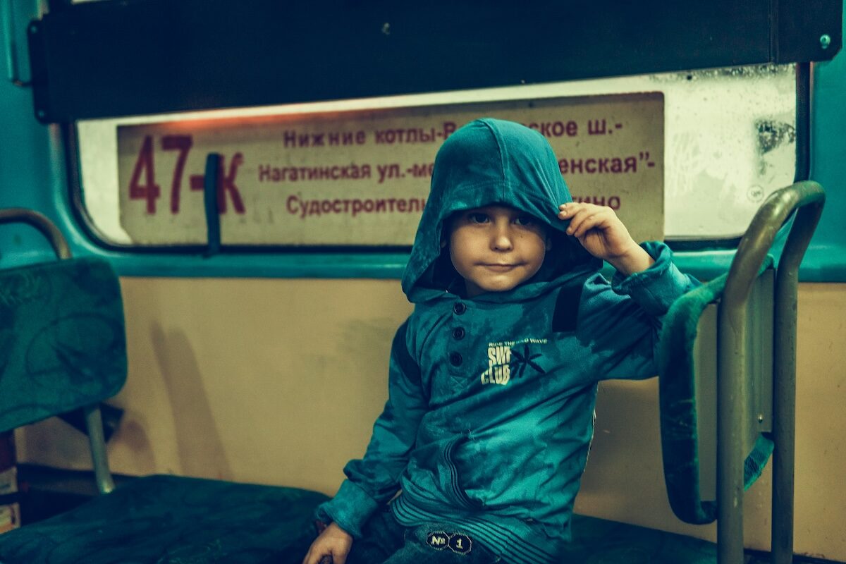 A young child in a blue jacket with a hood sits alone on a bus seat, partially covering their face with the hood. A sign with route 47-K is visible behind them.