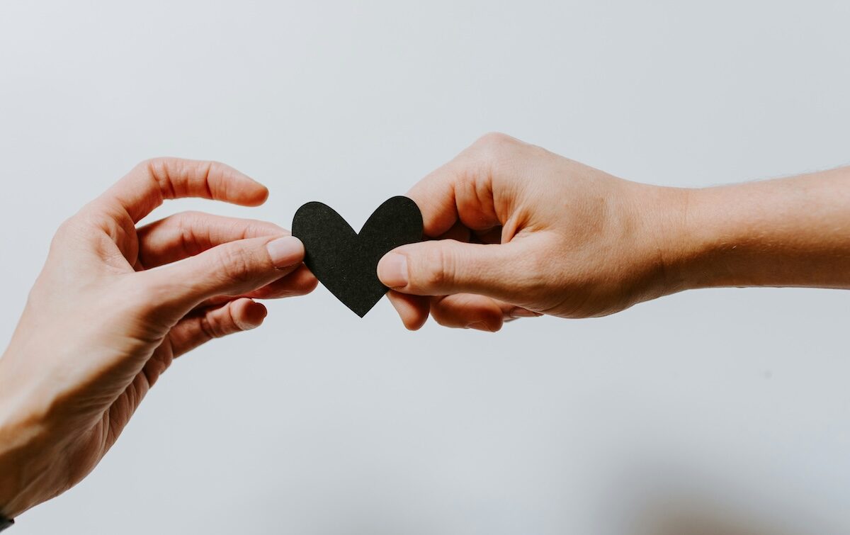 Two hands hold a small black paper heart between them against a plain white background.