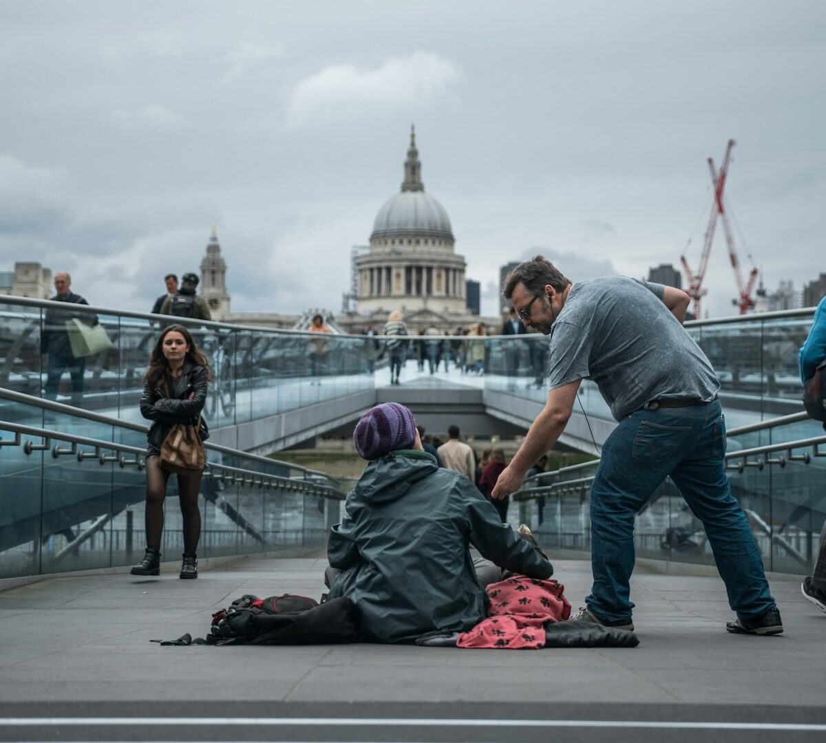 A man hands something to a seated person on a bridge, with St. Paul’s Cathedral and construction cranes visible in the background.