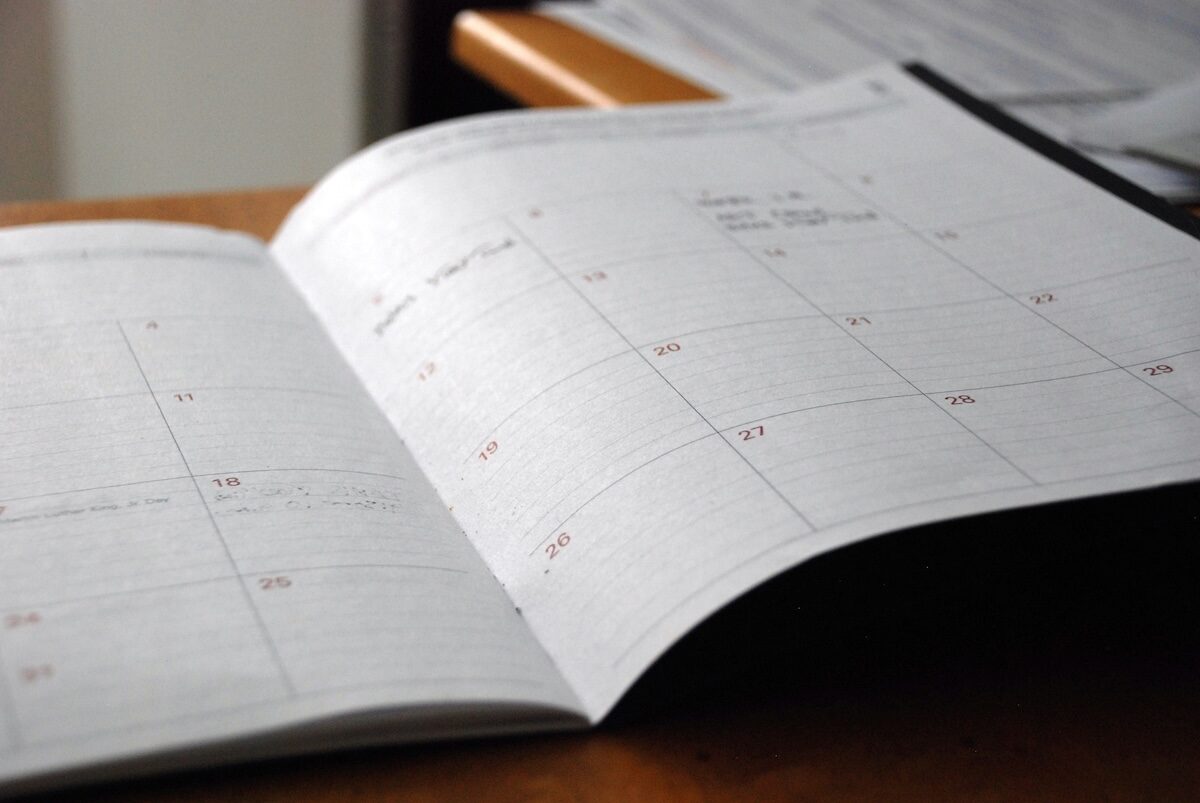 An open planner or calendar rests on a wooden desk, displaying a monthly layout with several dates and notes written in pen.