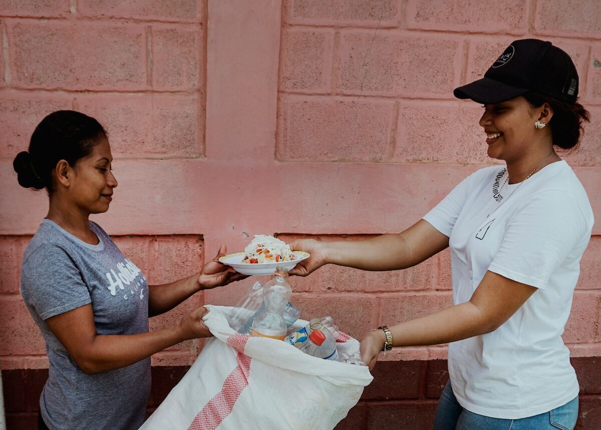 Two women stand in front of a pink wall; one hands a plate of food to the other, who is holding a large bag filled with items.