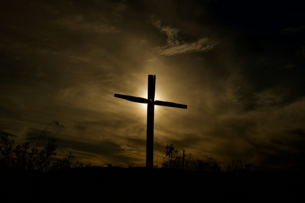 A wooden cross silhouetted against a dramatic, cloudy sky with the sun shining behind it.