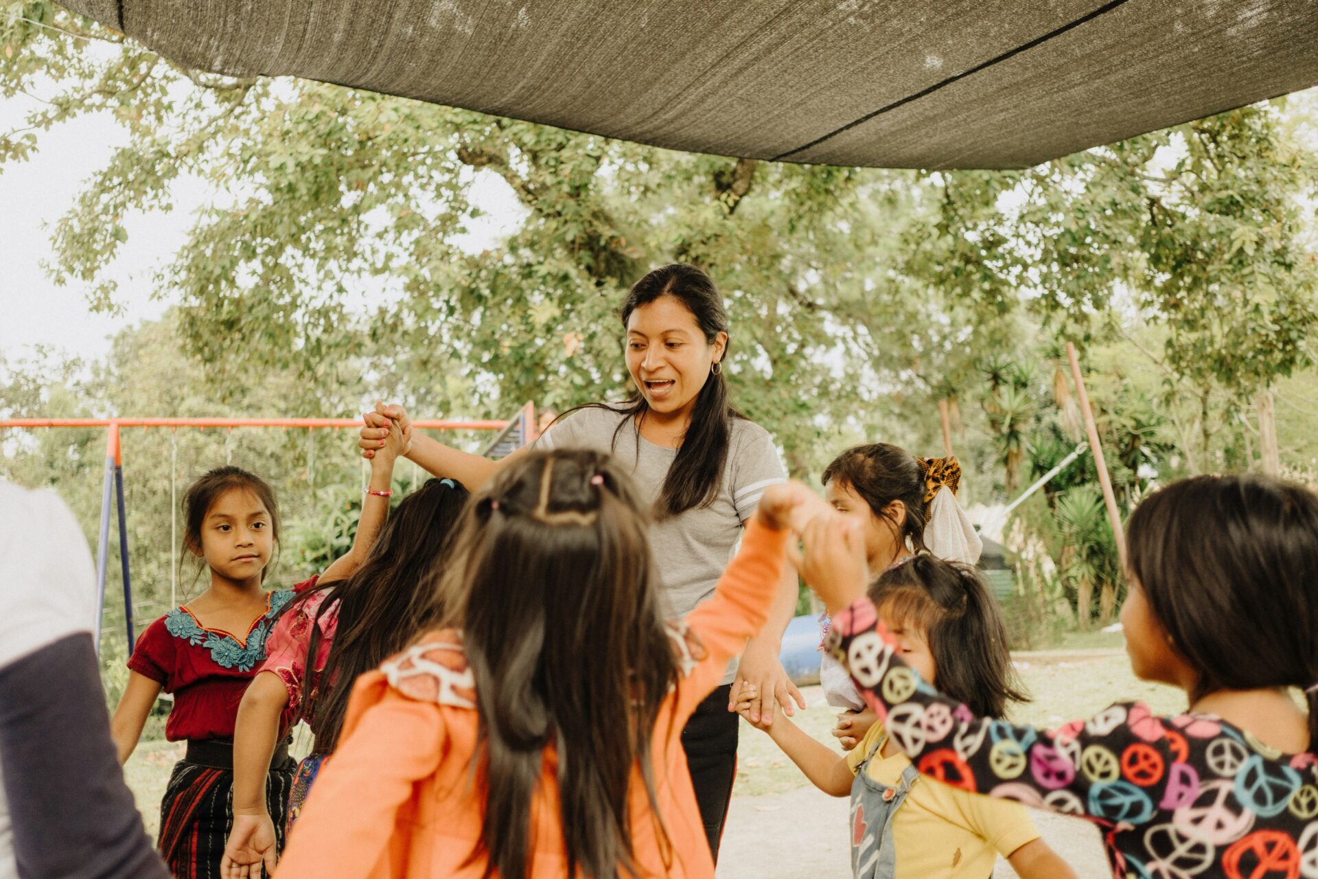 A woman and several children stand in a circle holding hands outdoors under a shade canopy, with trees and a swing set in the background.