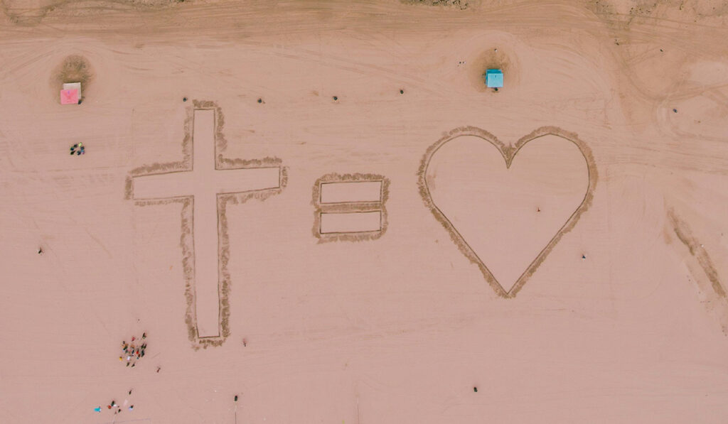 A cross, and equal symbol, and a heart are carved in the sand on a beach.