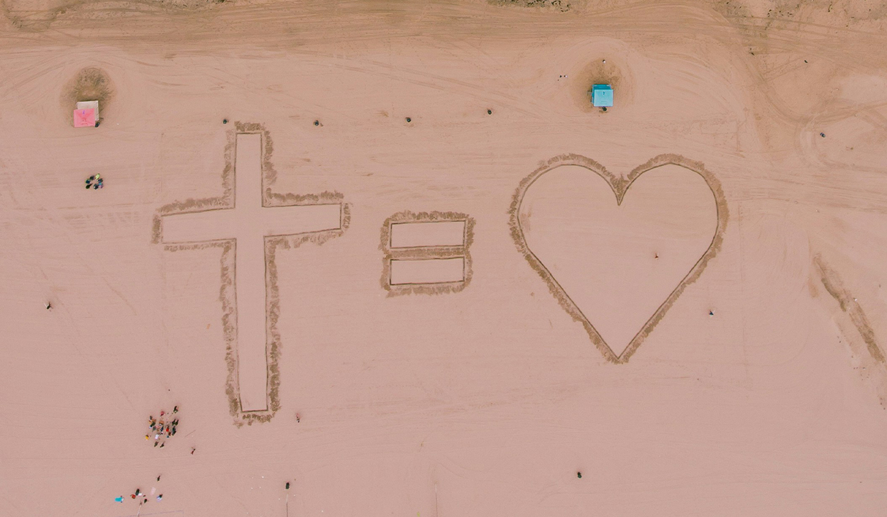 A cross, and equal symbol, and a heart are carved in the sand on a beach.
