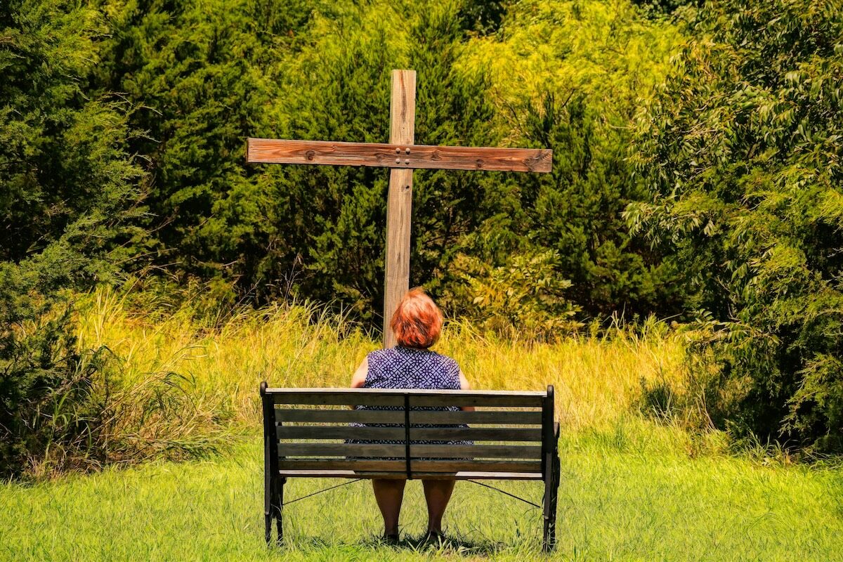 A person with short hair sits on a bench facing a large wooden cross in a grassy area surrounded by trees.