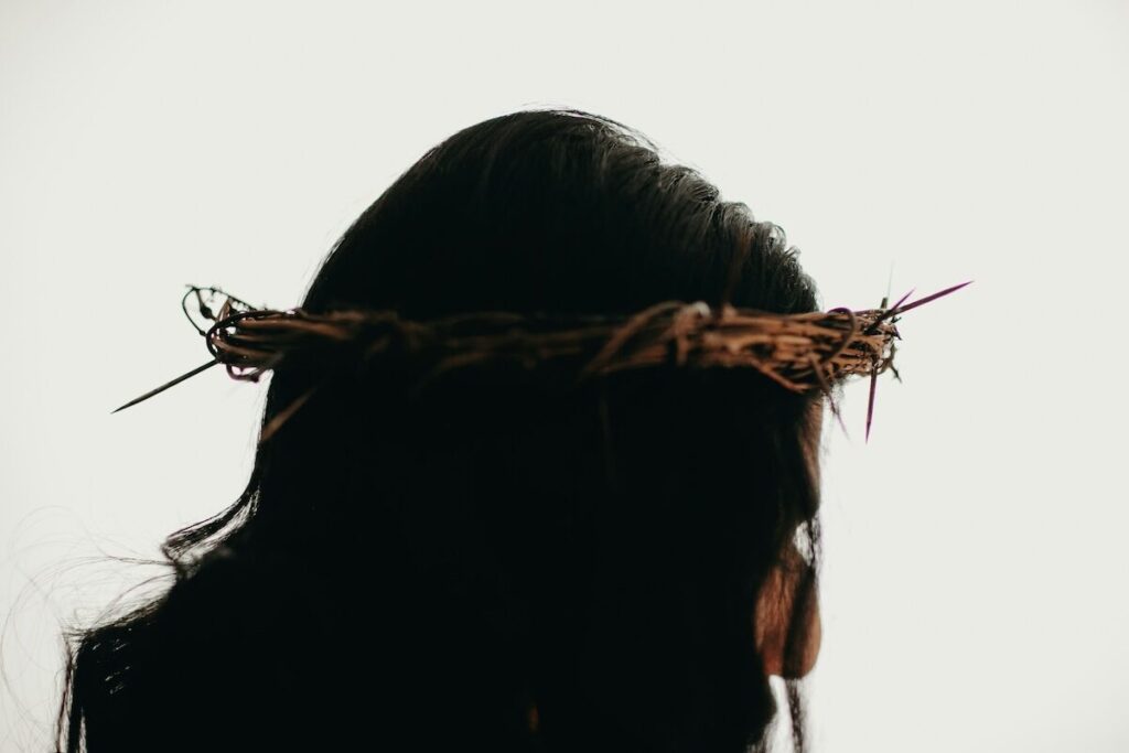 A person with long dark hair wearing a crown of thorns, shown from the back and side against a plain light background.