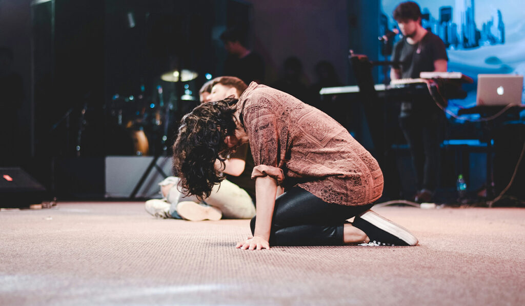 A woman on a stage is kneeling on the ground with her head hanging down. Her hair is covering her face.