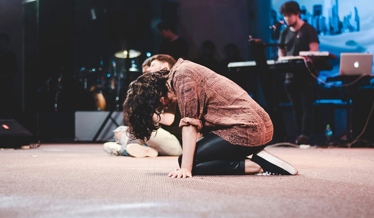 A woman on a stage is kneeling on the ground with her head hanging down. Her hair is covering her face.