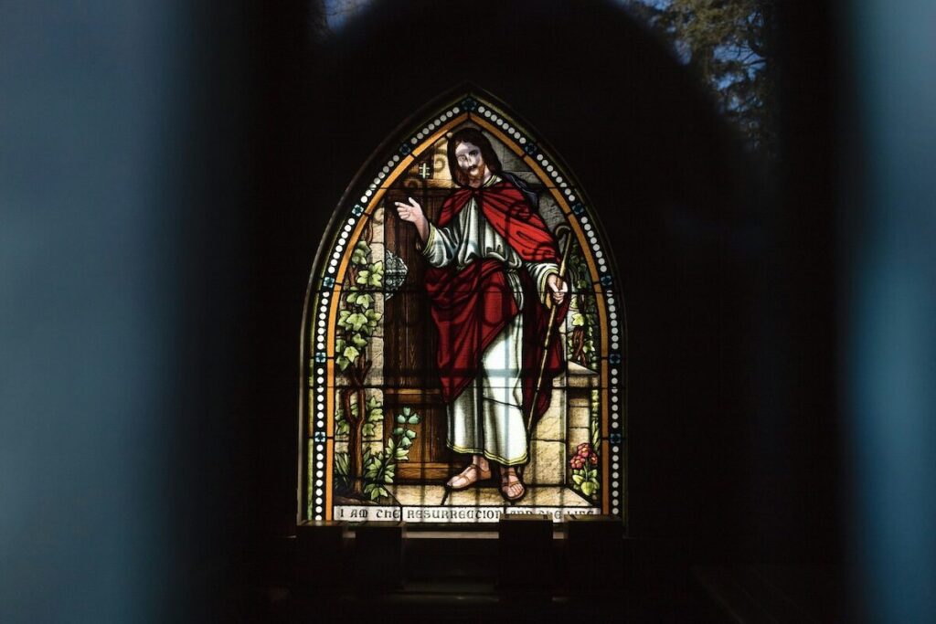 Stained glass window depicting a figure in white robes and a red cloak, standing with one hand raised, framed by an arched window.