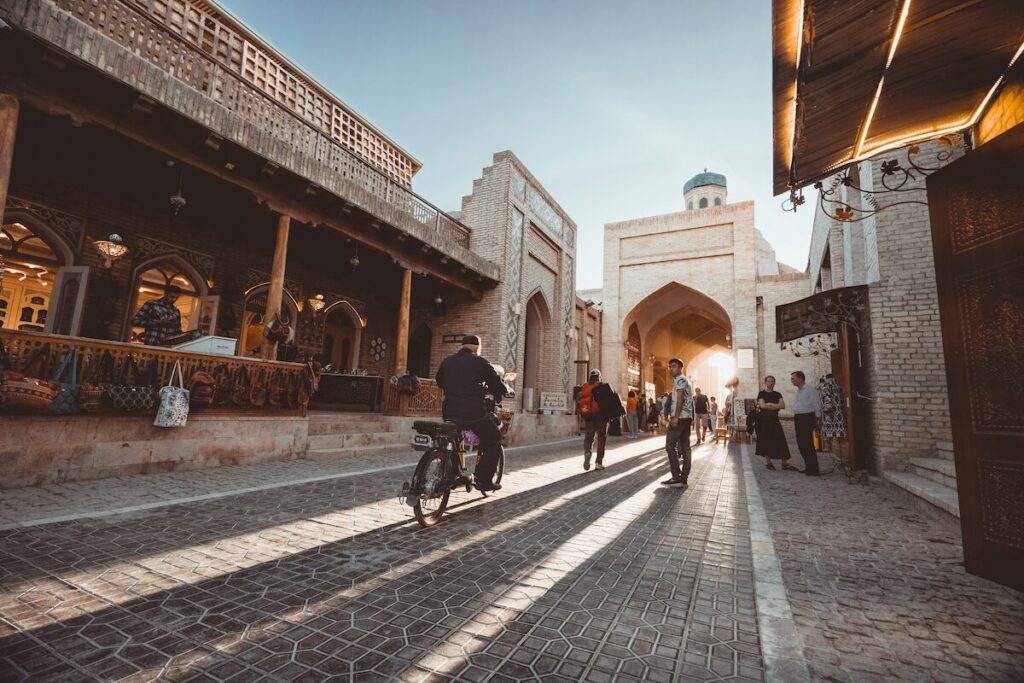 People walk and ride bicycles down a cobblestone street lined with historic buildings, leading toward an arched entrance in the background under a clear sky.