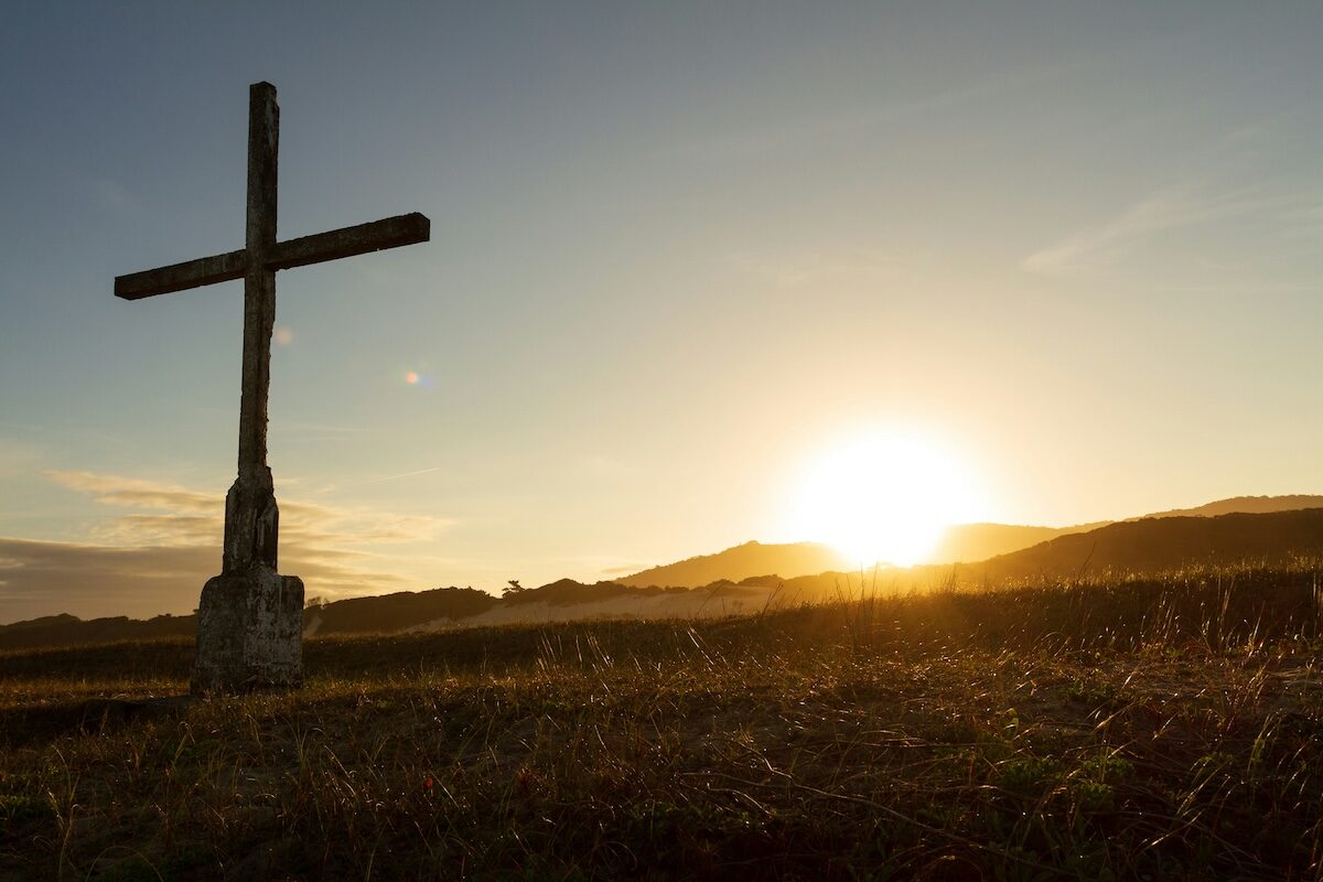 A wooden cross stands in a grassy field with the sun setting behind distant hills, casting a warm glow over the landscape.