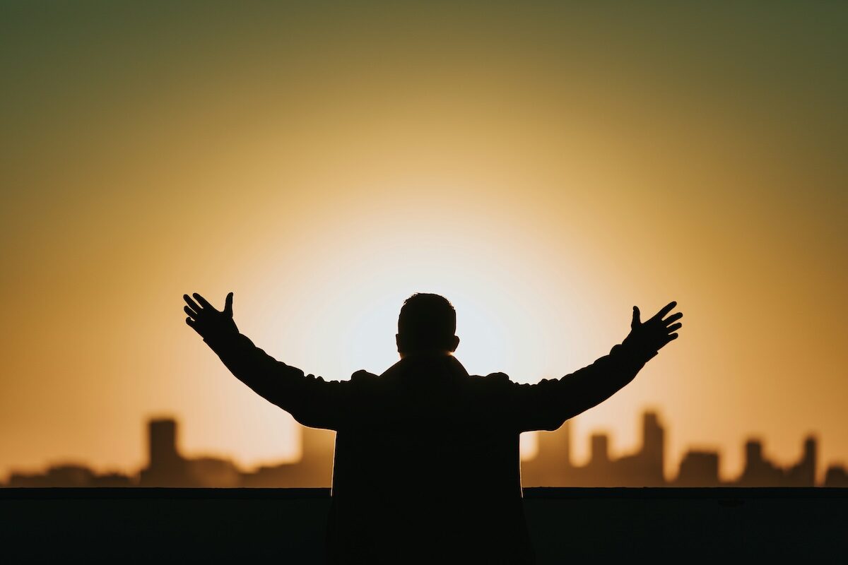 Silhouette of a person with arms outstretched facing a city skyline at sunrise or sunset.