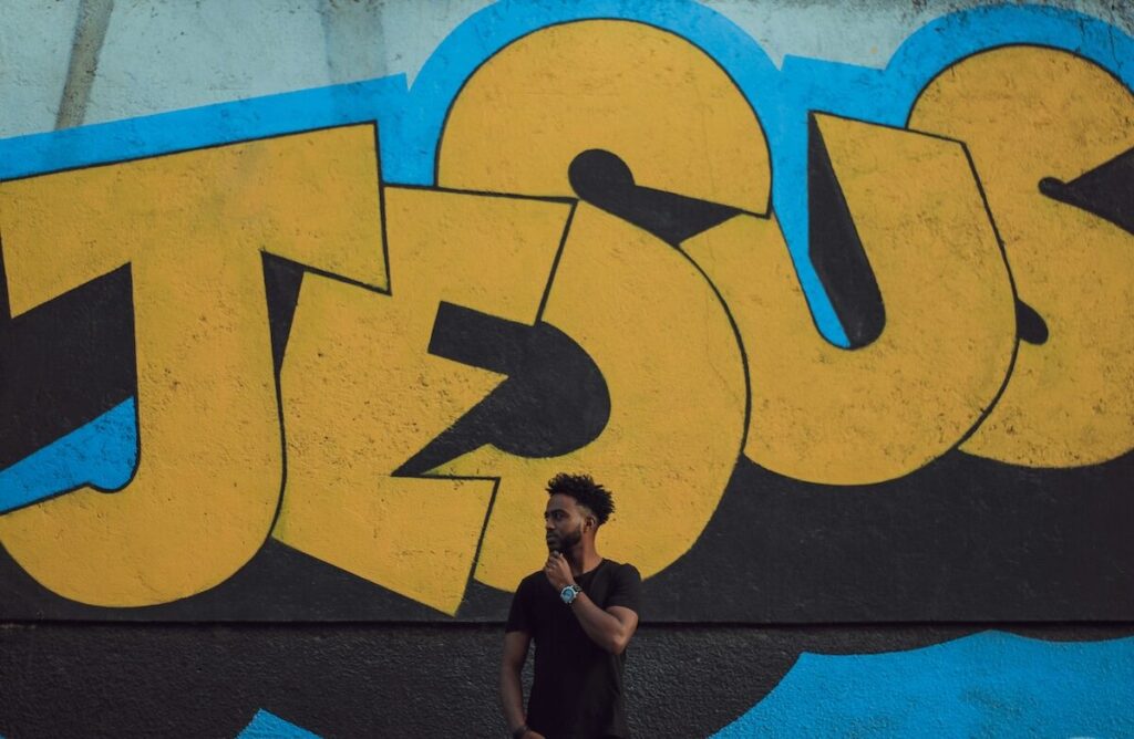 A man stands in front of a mural with large yellow letters spelling JESUS on a blue and black background.